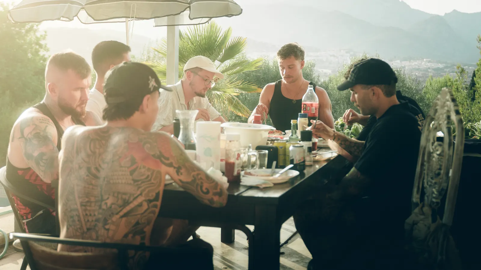Eine Gruppe Männer isst gemeinsam an einem langen Tisch auf der Terrasse, dahinter die Berge.