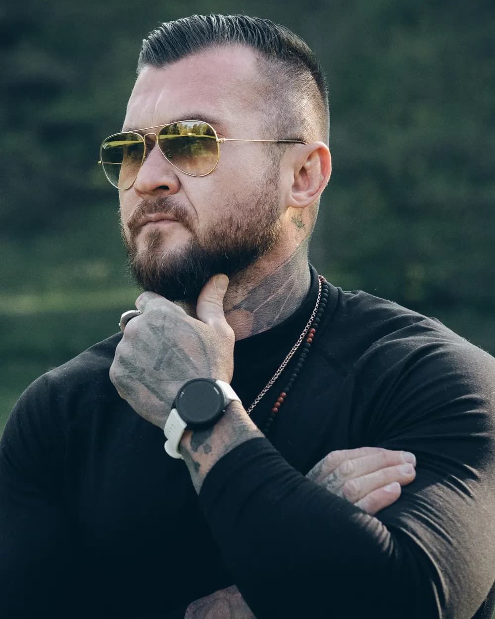 Jurij Sinenkov im schwarzen Longsleeve mit Sonnenbrille auf einer Waldlichtung, die Hand am Kinn.