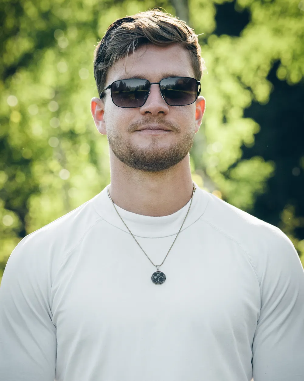 Timo im weißen Longsleeve mit Sonnenbrille vor sonnigem Grün.